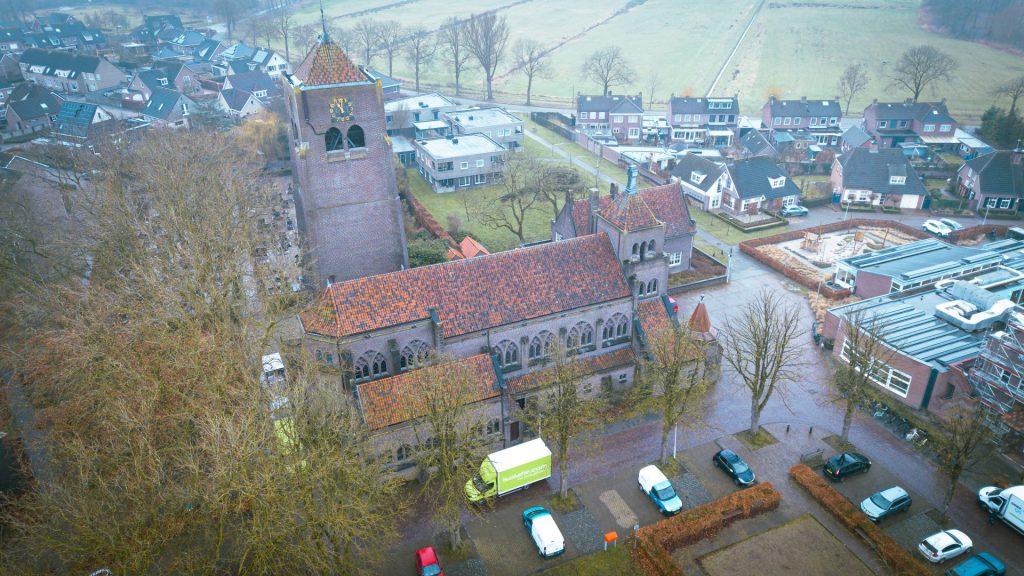 Een overzicht van een dorp met een oude kerk, omringd door huizen en bomen, op een regenachtige dag, met parkeergelegenheid voor auto's.